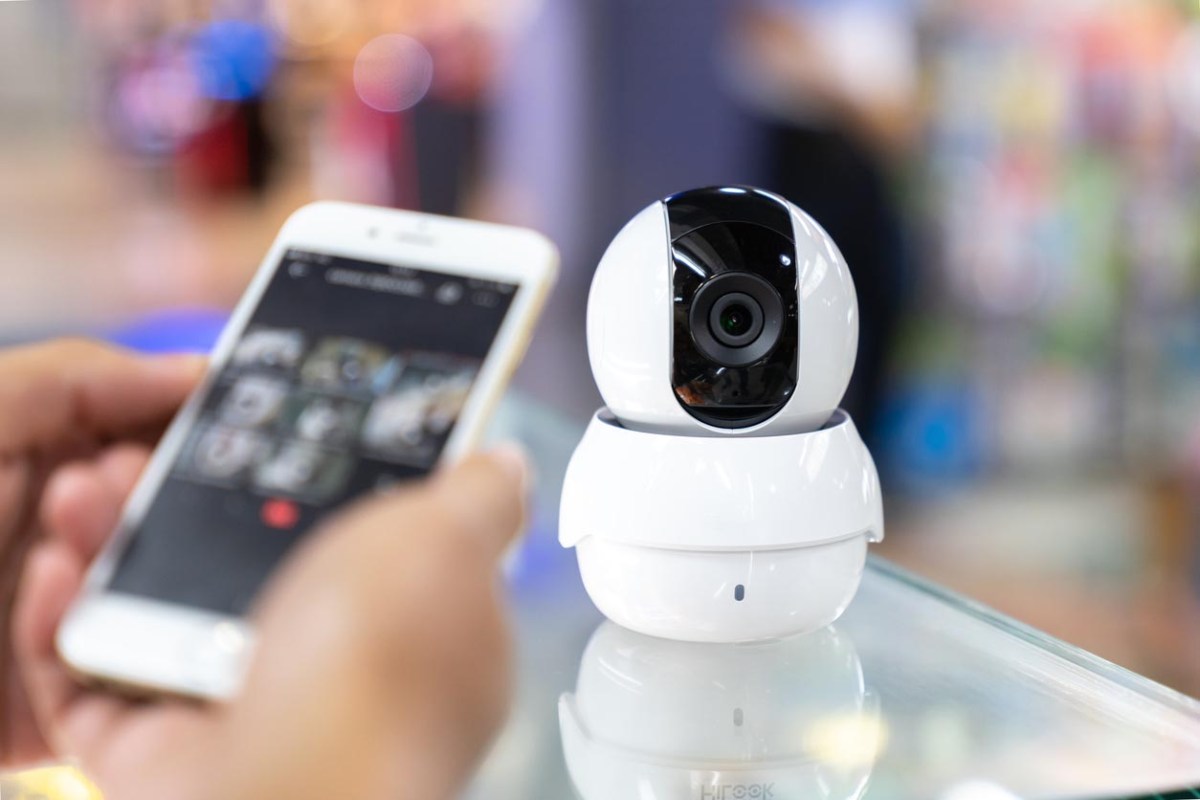 A home security camera is featured front and center, with a smart phone screen in the foreground.