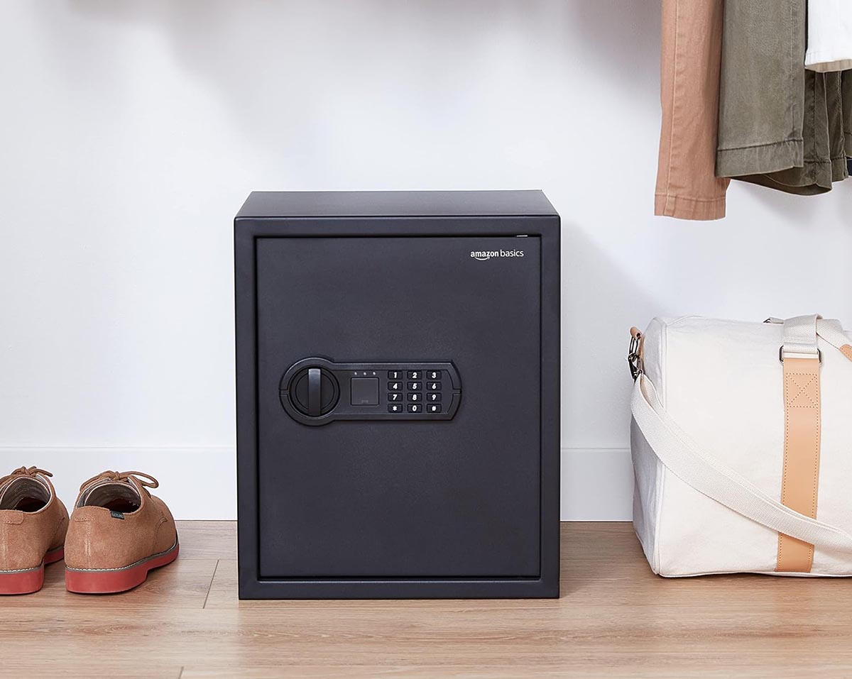 A close up of an Amazon Basics safe on the floor between a duffle bag and a pair of shoes. 