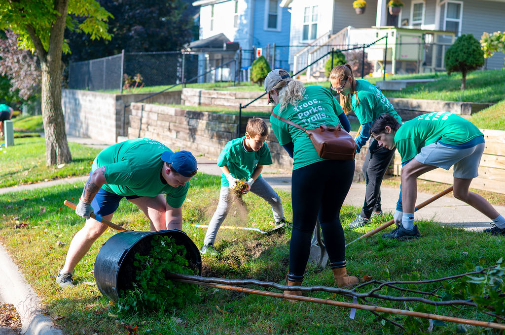A multigenerational group of people wearing matching green t-shirts work together to plant a tree.