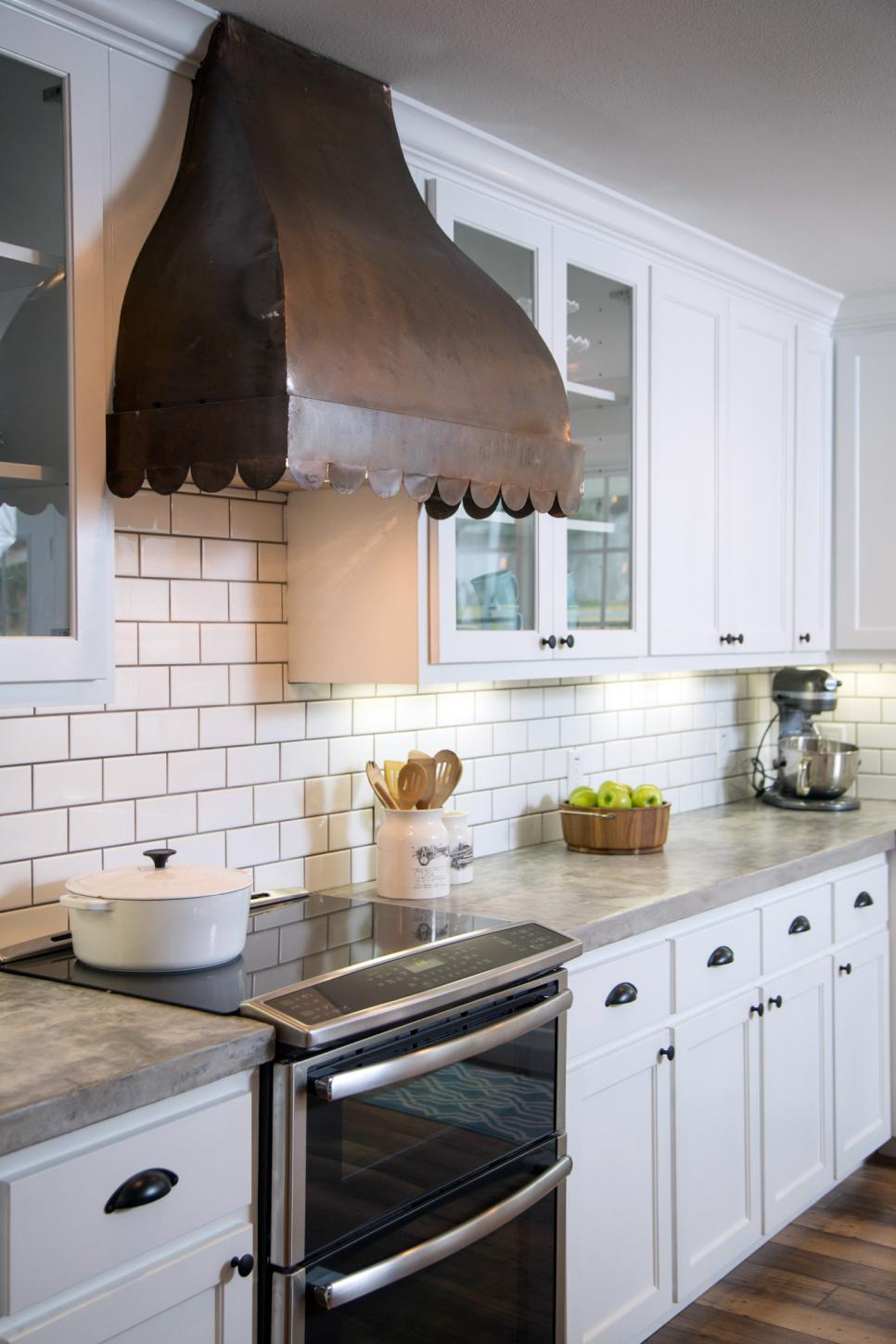 A rustic range hood with scalloped trim in kitchen with white cabinets and white tile backsplash.