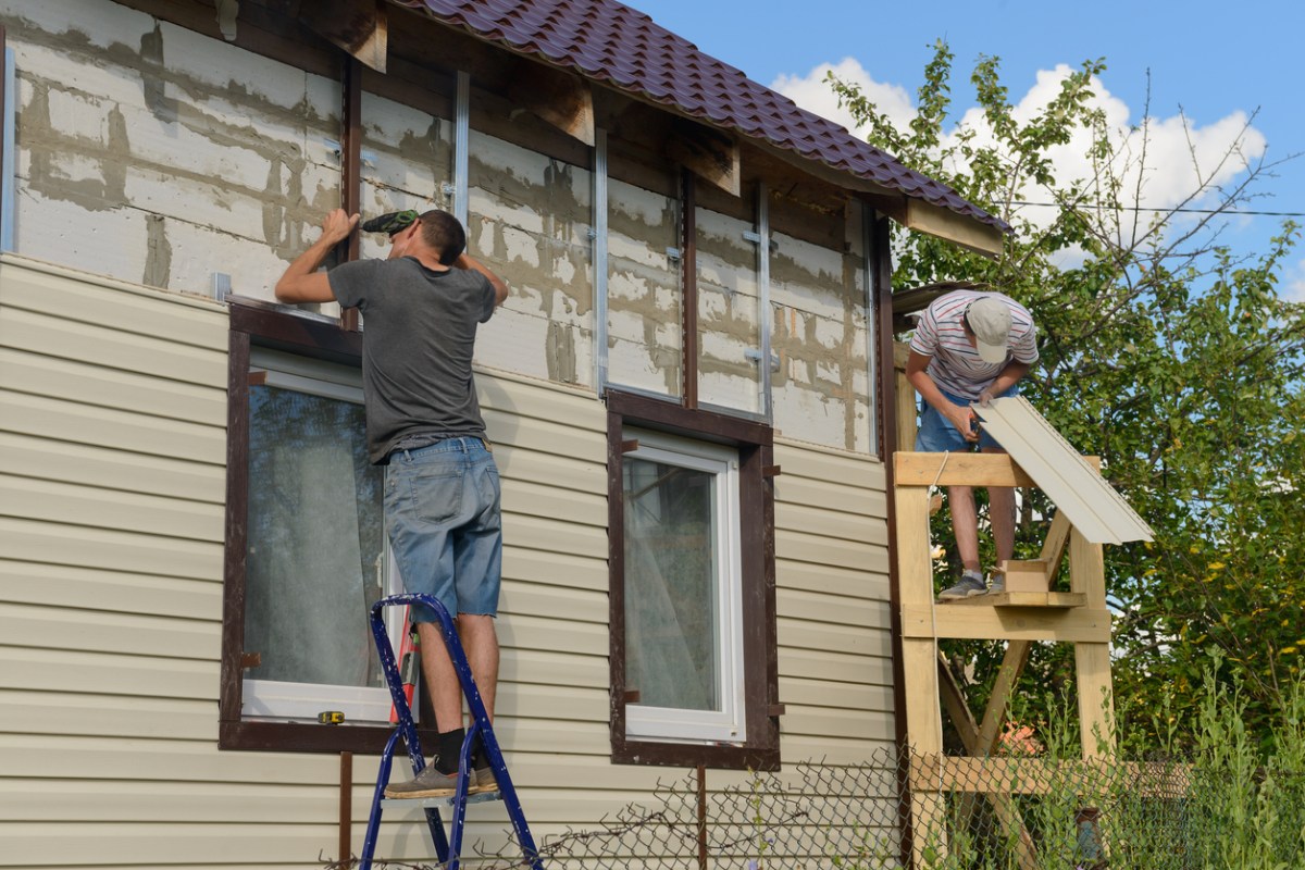 Two workers install vinyl siding on a house on a bright and sunny day.