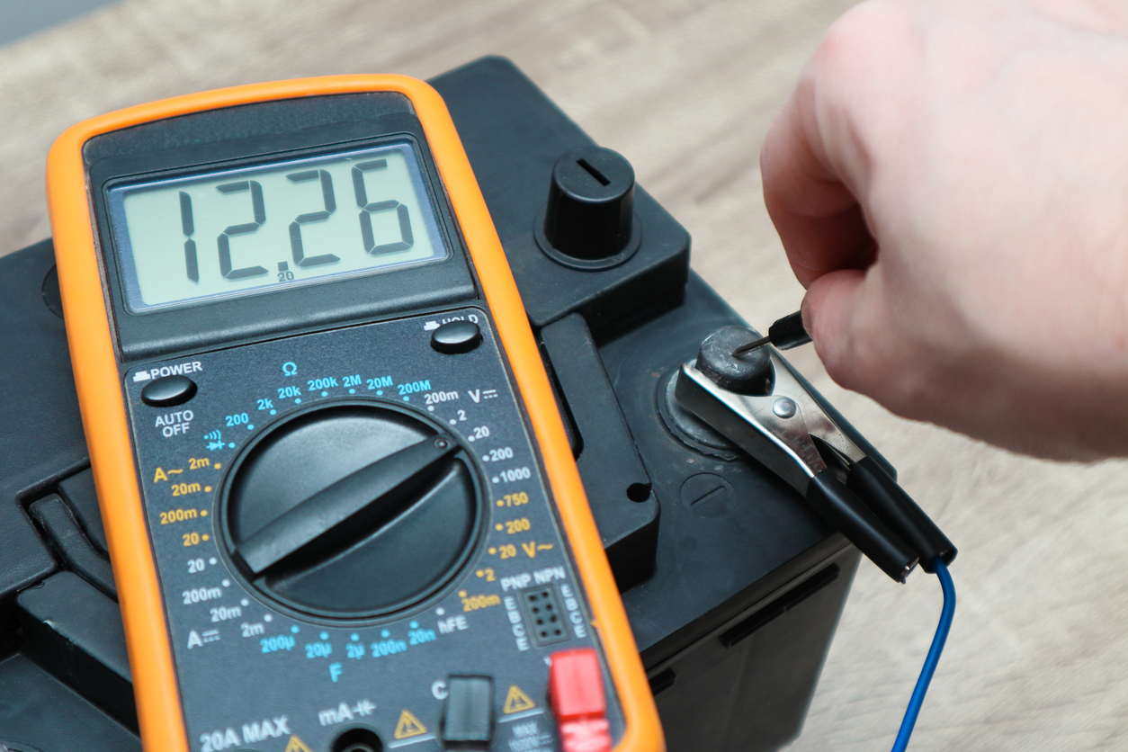 A person touches the needle of a multimeter to the negative terminal of a car battery for a reading of 12.26 volts.