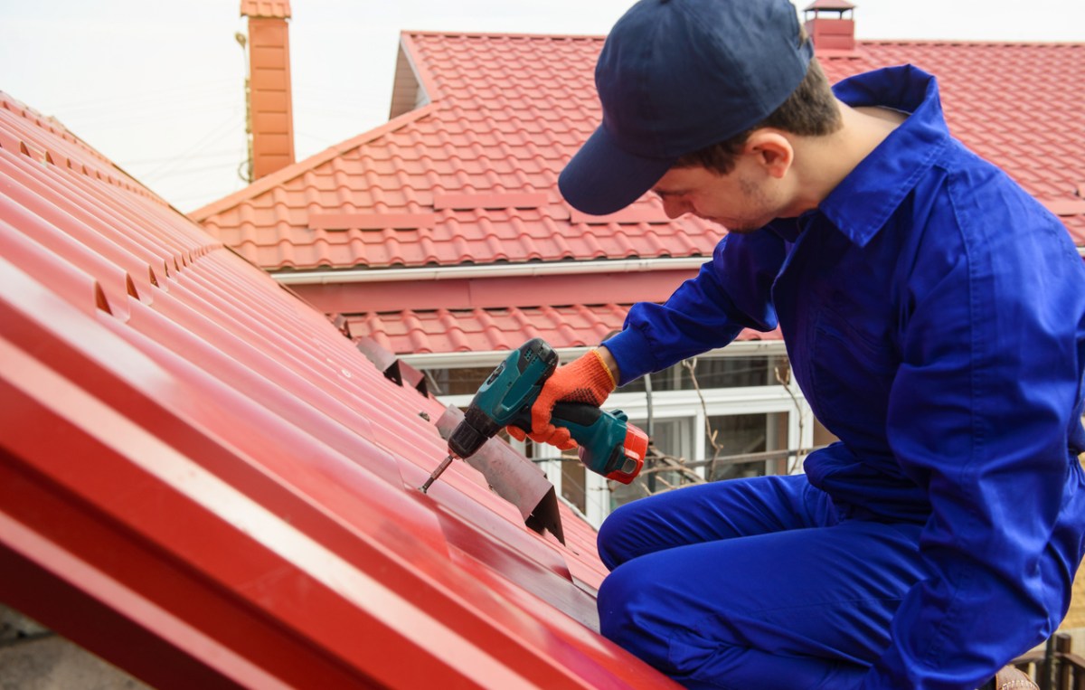 A man in a blue cap and a blue work suit uses a drill to install a red roof. 