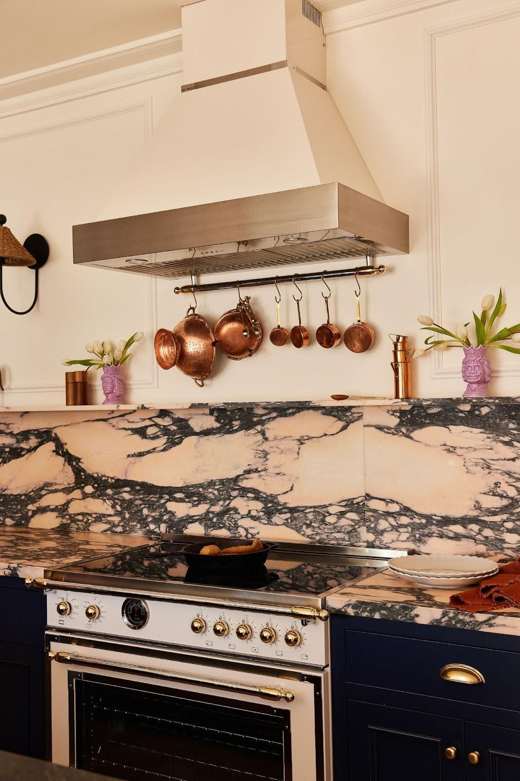 A white range hood with stainless steel trim with marble backsplash and copper pots.