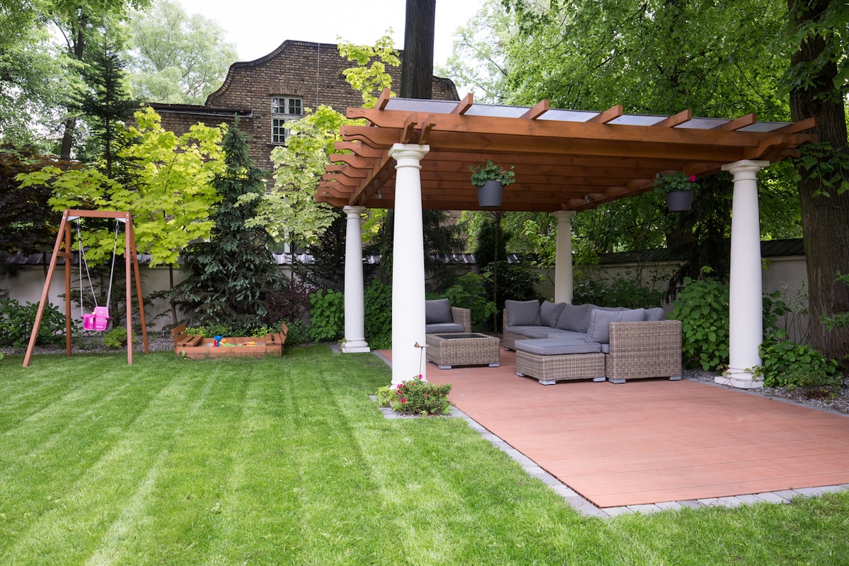 A fenced-in backyard with a paved outdoor living area and lawn furniture, which is covered by a pergola.