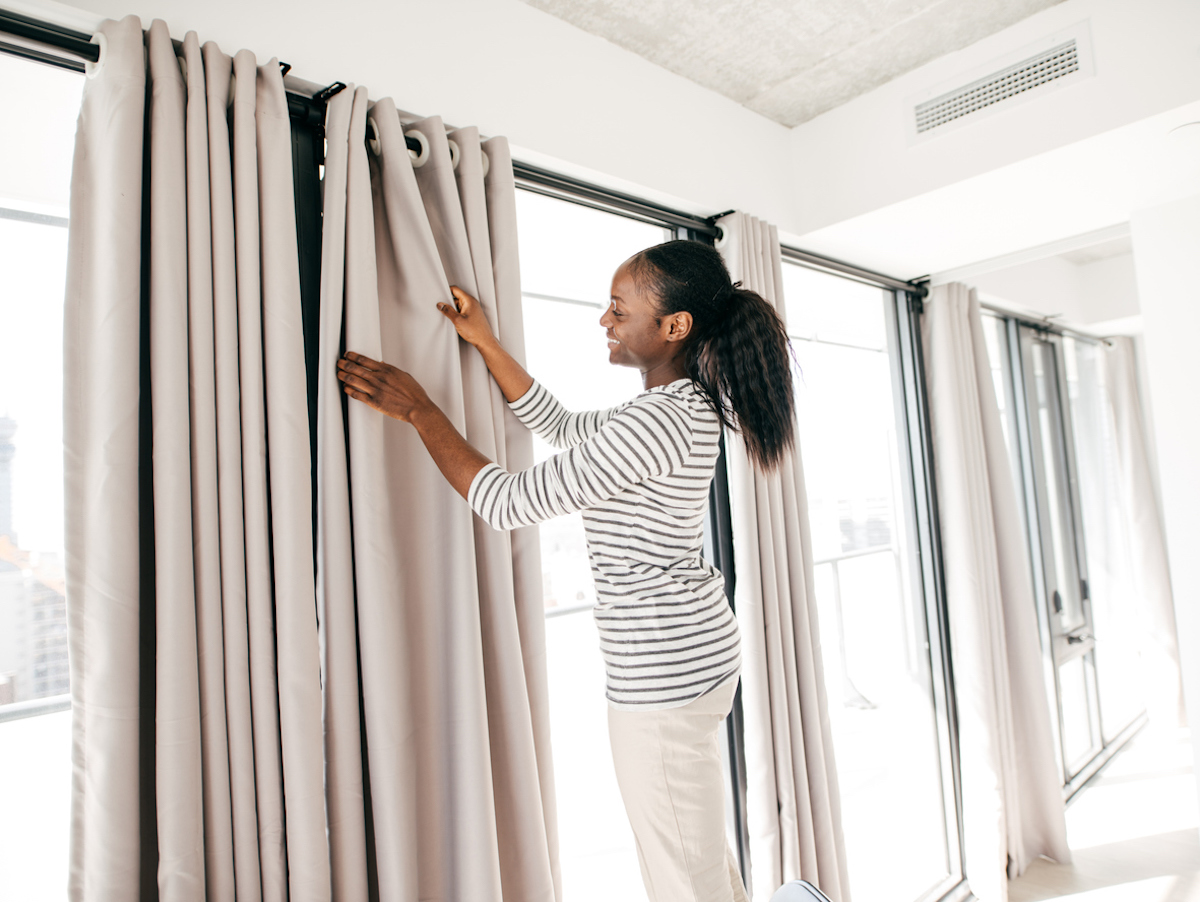 Woman on step stool hanging beige curtains over a wall of windows.
