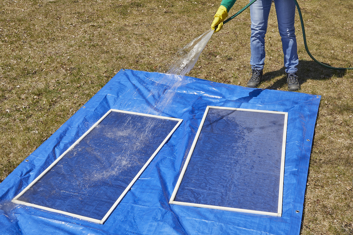 Two window screens on a blue plastic tarp being rinsed by a woman holding a garden hose.