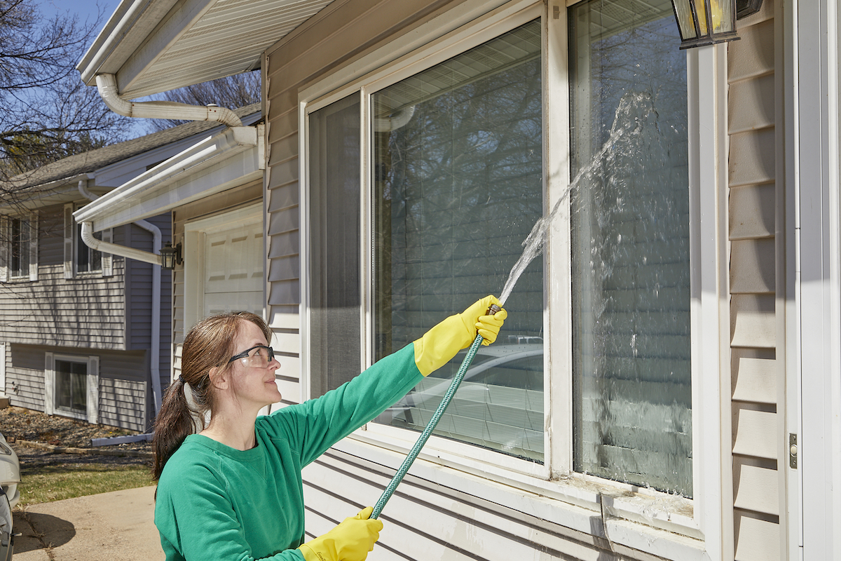 Woman wearing a green shirt and rubber gloves uses a garden hose to rinse exterior windows.