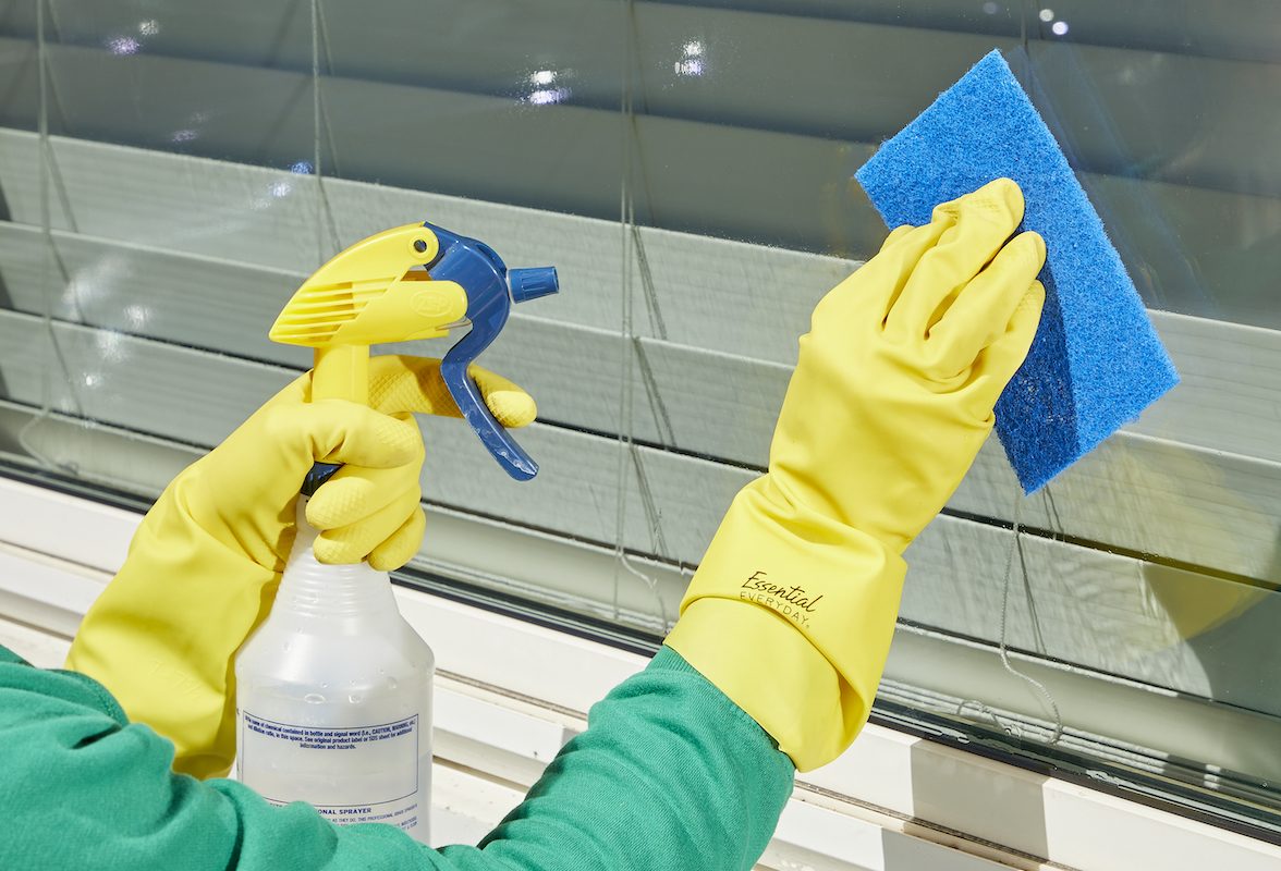 Woman cleaning an exterior window with a spray bottle and a blue scrub sponge.