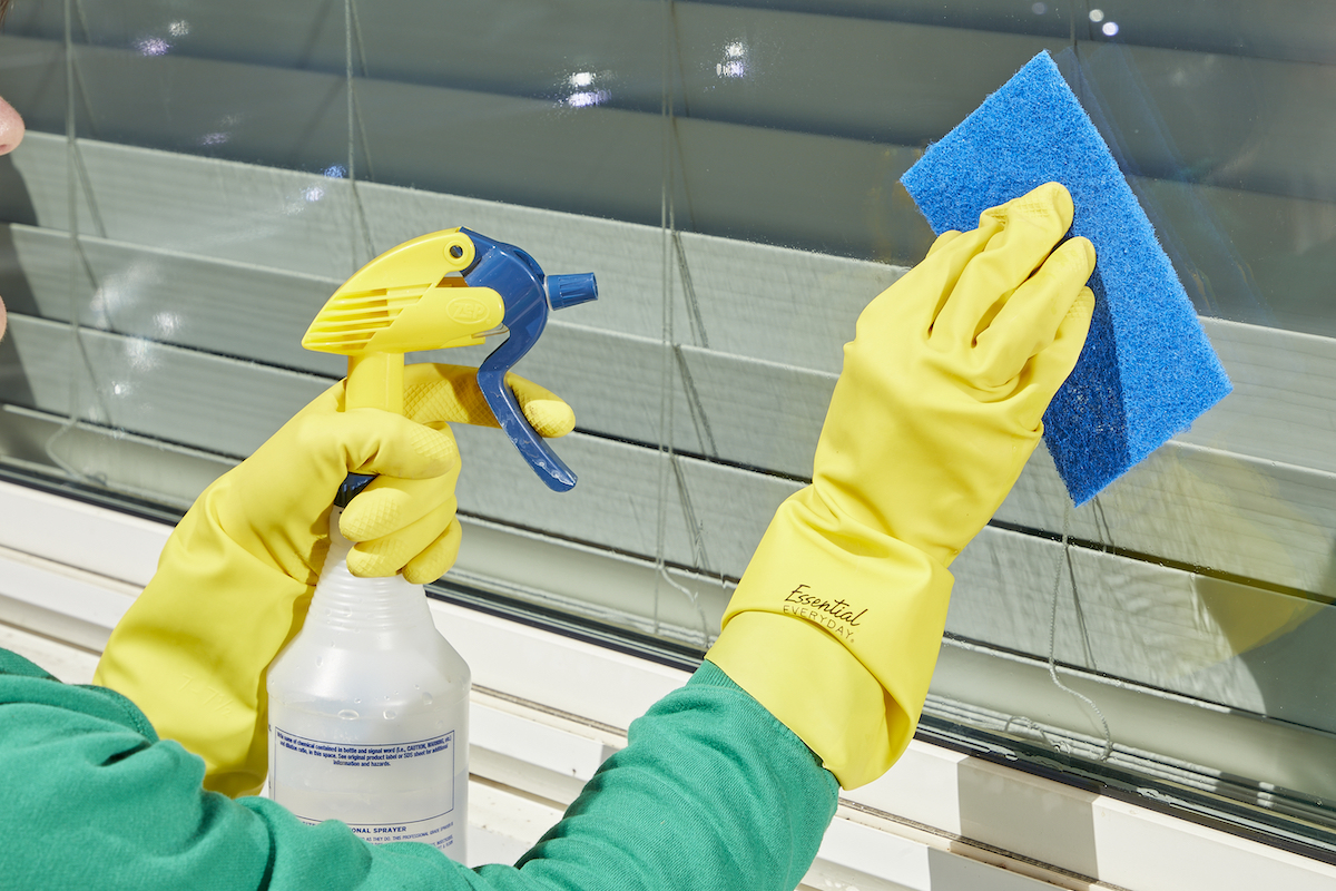 Woman cleaning an exterior window with a spray bottle and a blue scrub sponge.