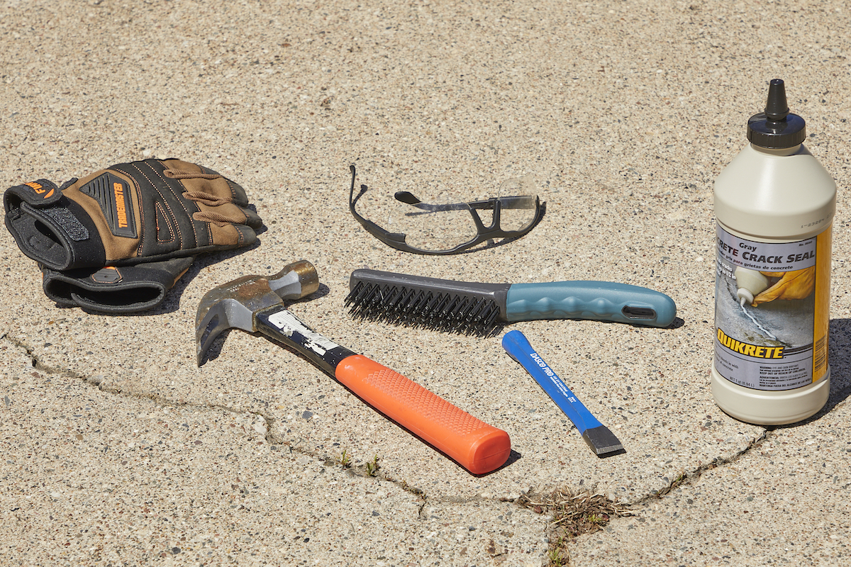 Materials needed to fix cracks in concrete—hammer, wire brush, safety glasses and more—laid out on a concrete driveway.