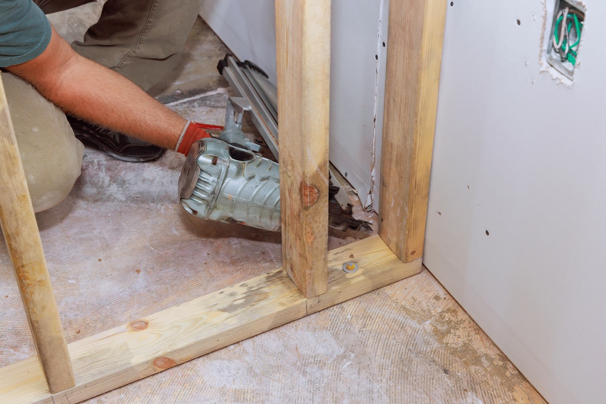 A person nailing studs to a bottom plate installed in an unfinished basement.