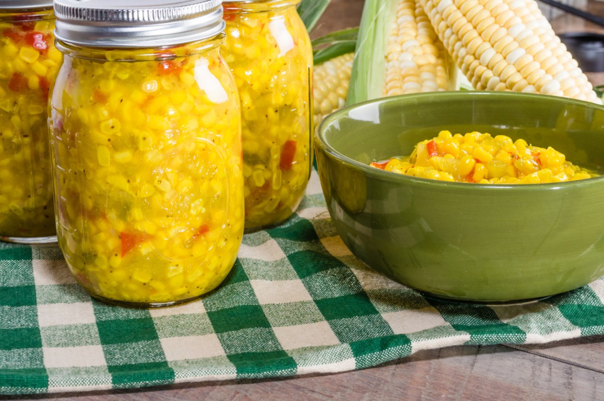Corn relish jar next to serving bowl on plaid table cloth.