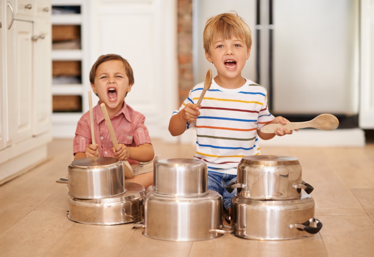 Two young boys sitting on the floor, using kitchen spoons to bang on pots and pans.