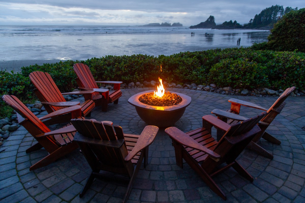 Red Adirondack chairs are positioned in a circle around a firepit on a lakeside patio.