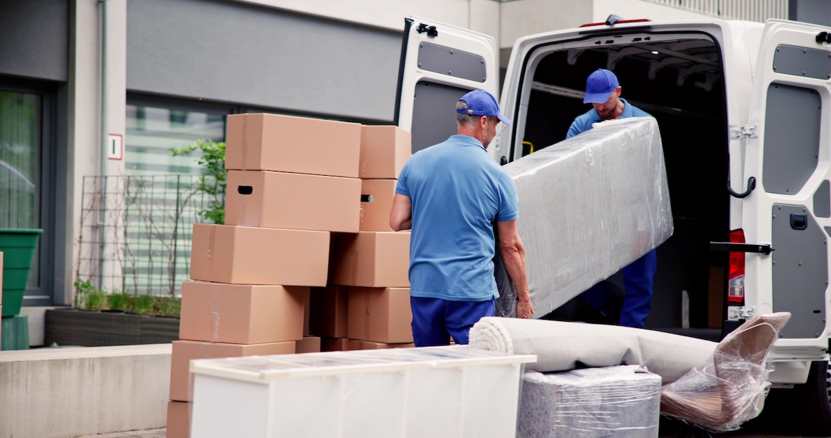 Two professional movers in blue uniforms move boxes and mattress into a truck.