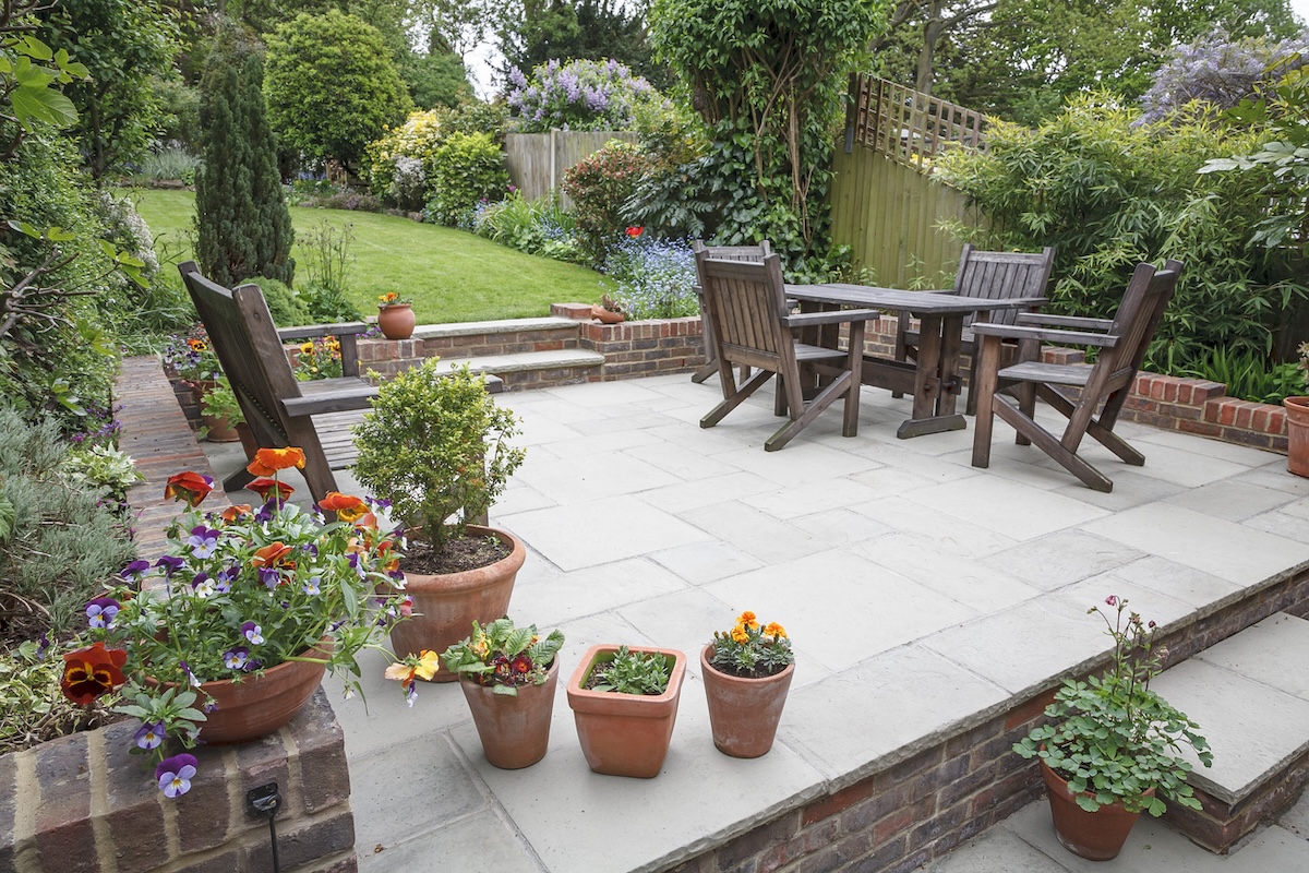 A hardscaped backyard with a stone and brick patio, a wooden table and chair set, and potted flowers.