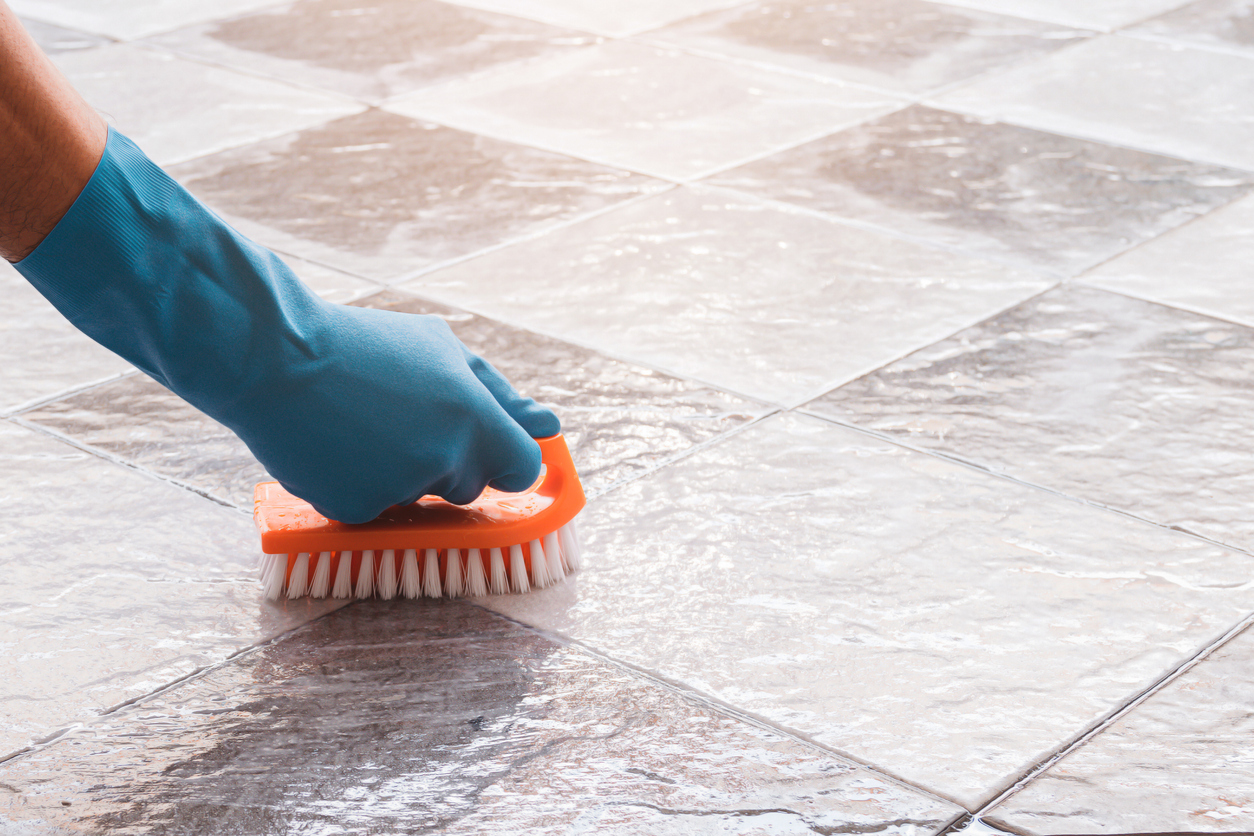 A person wearing blue latex gloves scrubs the grout between floor tiles with a stiff-bristled brush.
