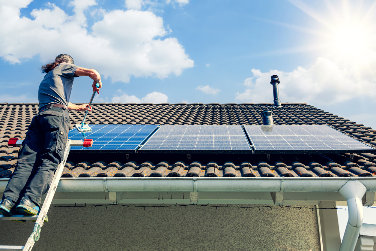 A person standing near the top of a ladder uses a Swiffer-like telescoping brush to clean the solar panels on a house roof.