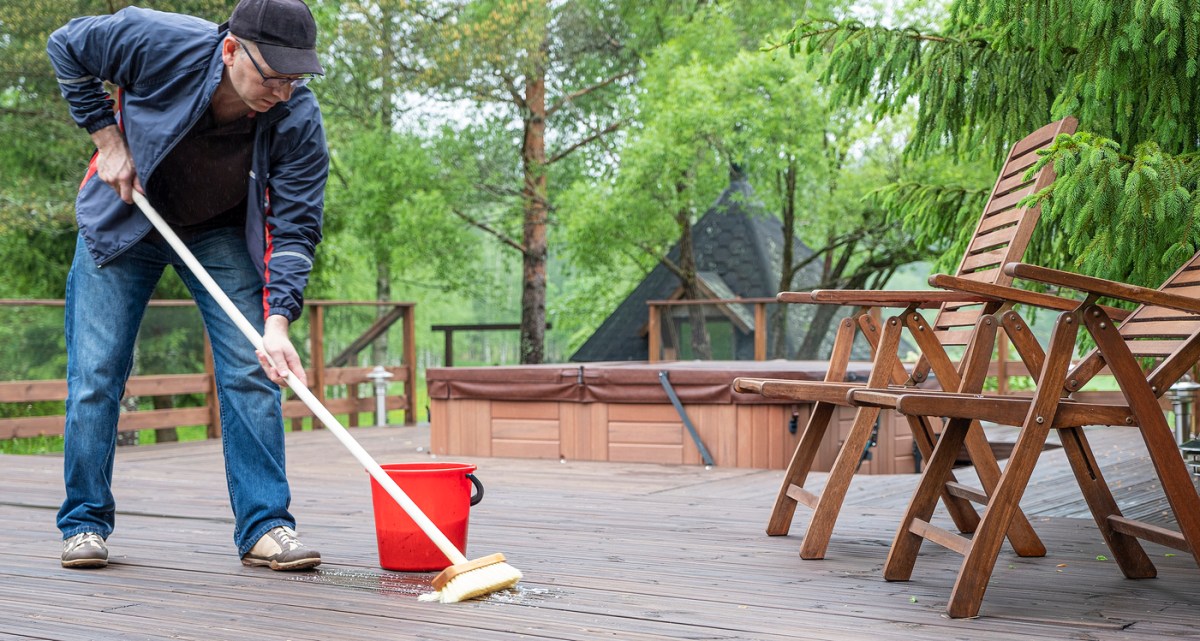 A mature person is bent over while scrubbing the surface of a large wooden deck with a brush. A red bucket with liquid inside sits directly beside him.