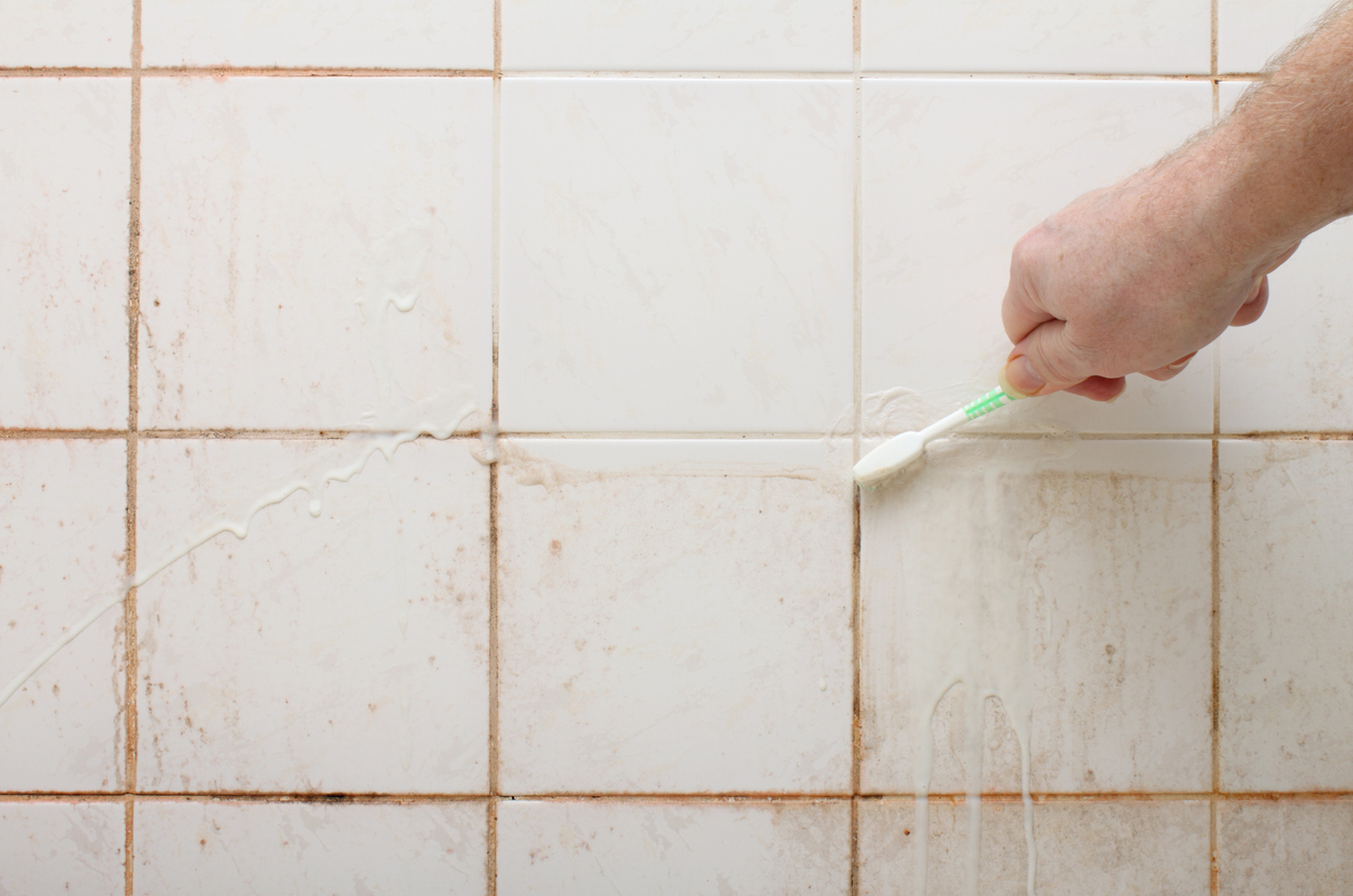 A person using a toothbrush to clean the grout between dirty shower wall tiles.