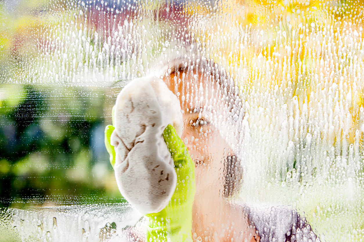 A young woman wearing yellow cleaning gloves cleans the exterior of a large window with a cloth.