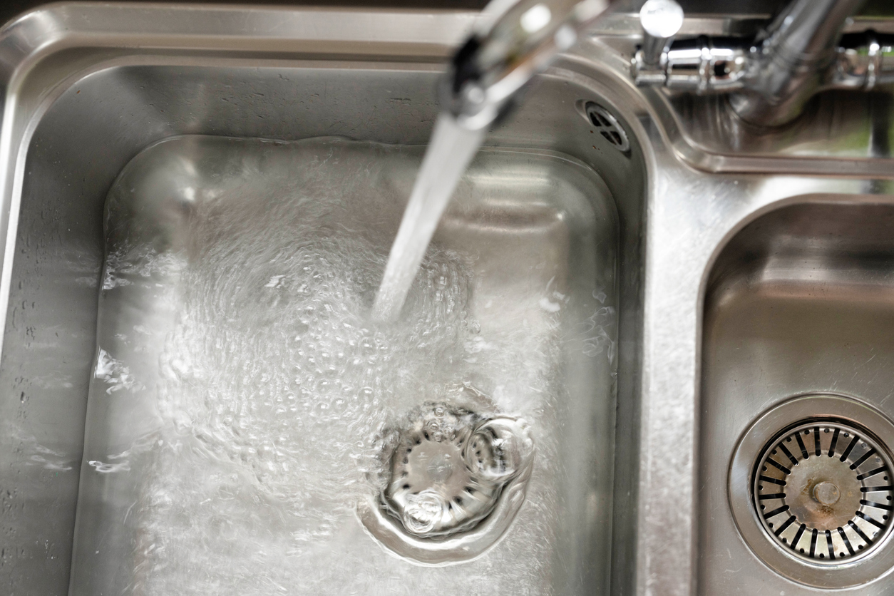 One basin of two in a stainless steel kitchen sink begins to fill with water from its running faucet.