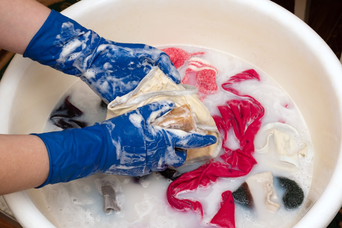 A person wearing blue gloves scrubs an article of clothing while more clothes soak in a sudsy basin.