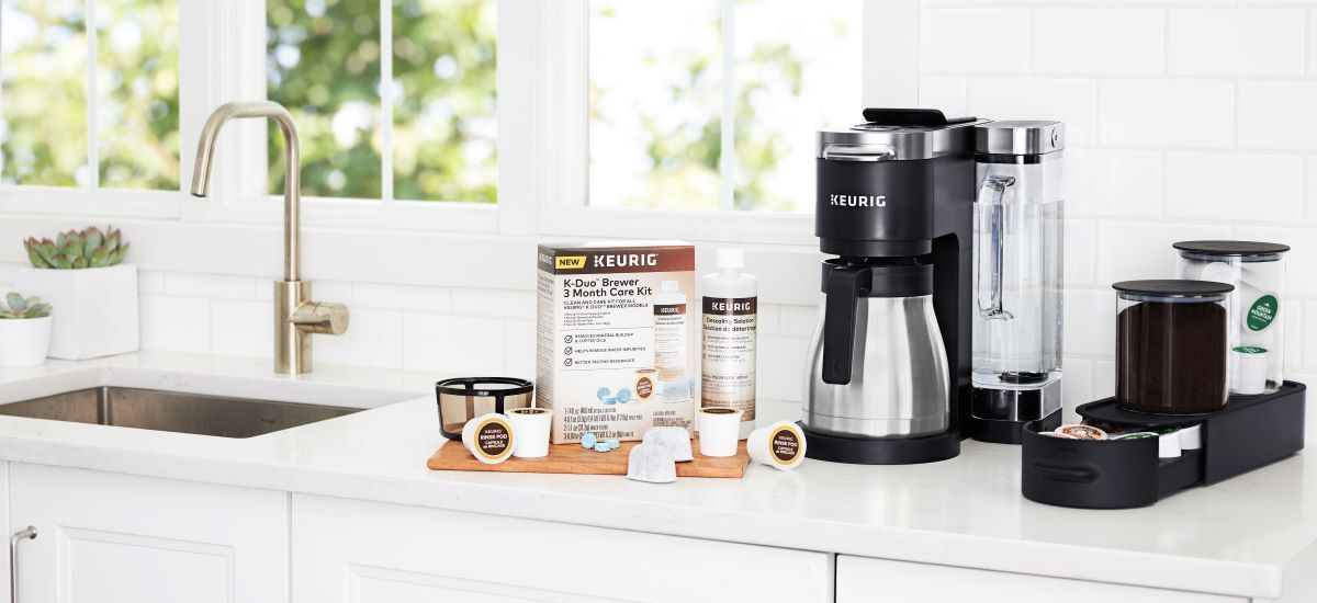 A Keurig K-Duo Brewer 3-Month Care Kit sits unboxed on a white kitchen counter next to a stainless steel and black Keurig coffee maker.