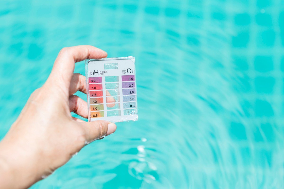 A person holds a small pool water test kit with a colored chart for pH balance and chlorine level readings printed on the front of it.