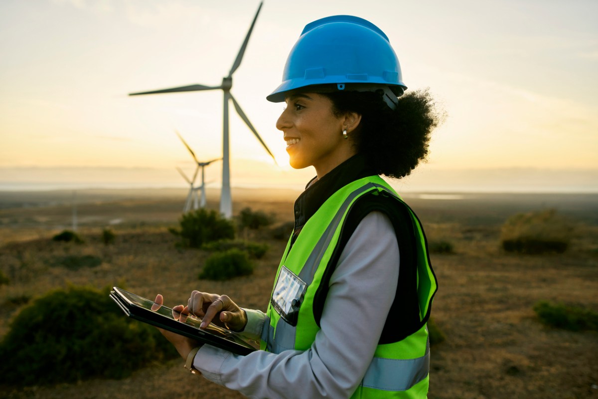A worker in a blue hard hat smiles as wind turbines spin behind her. 
