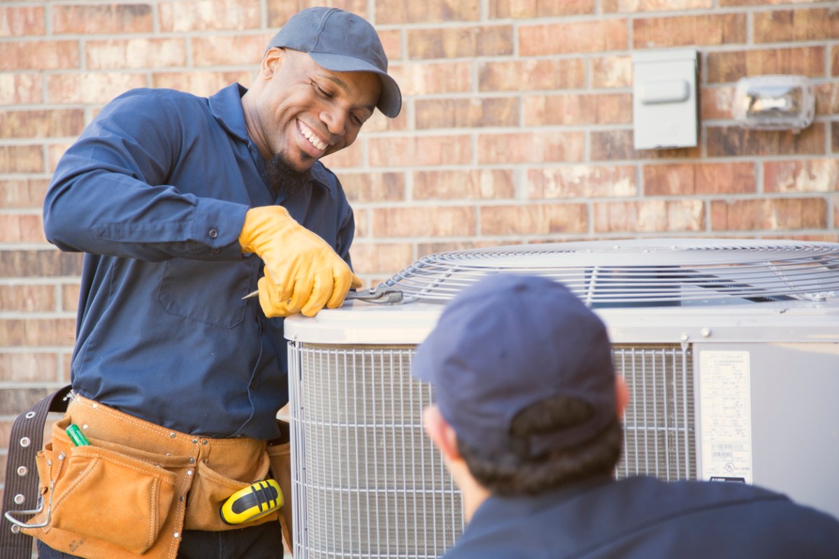 Two workers in blue shirts and caps fix an outside AC unit. 