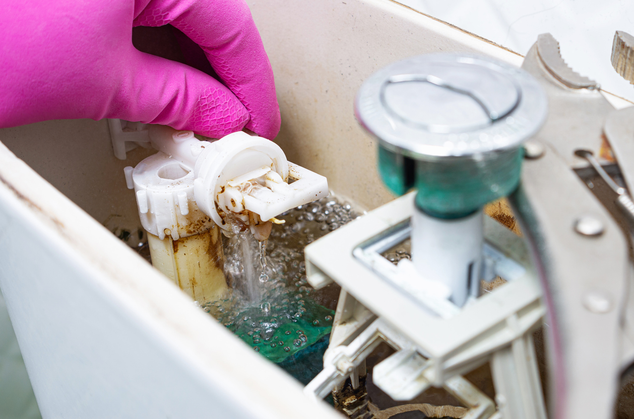 A pink-gloved hand reaches inside of an open toilet tank, which has signs of significant scale and possible mold buildup.