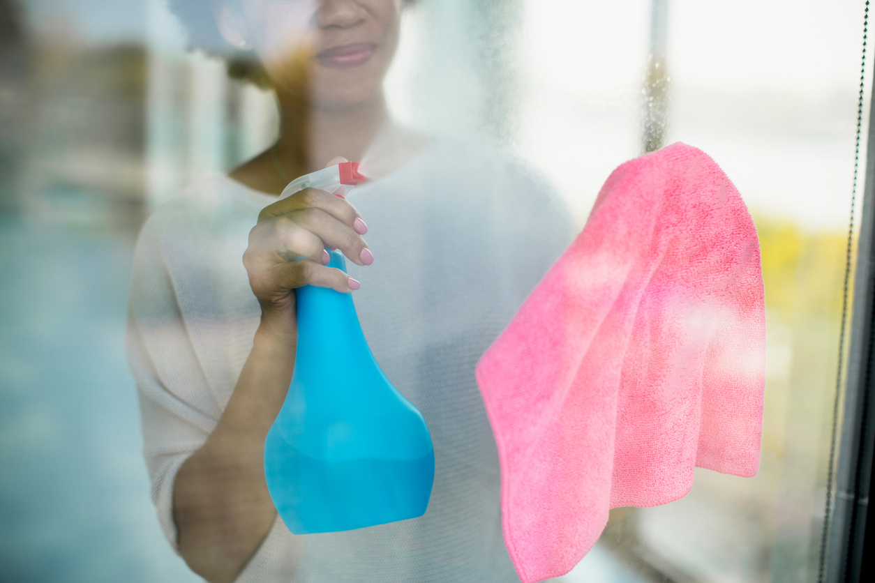 A woman standing behind a large glass window pane cleaning  and cleaning it with a pink microfiber cloth and homemade glass cleaner.