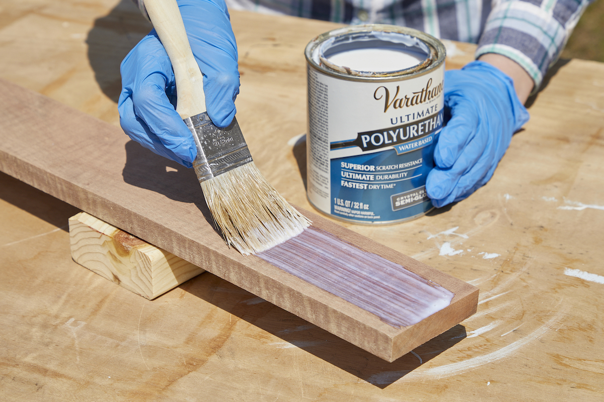 Woman applies polyurethane with a paintbrush to a piece of wood.