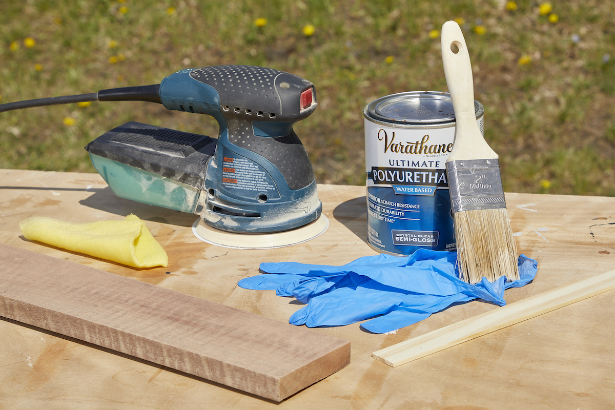 Palm sander, polyurethane, paintbrush and other materials laid out on a table outdoors.