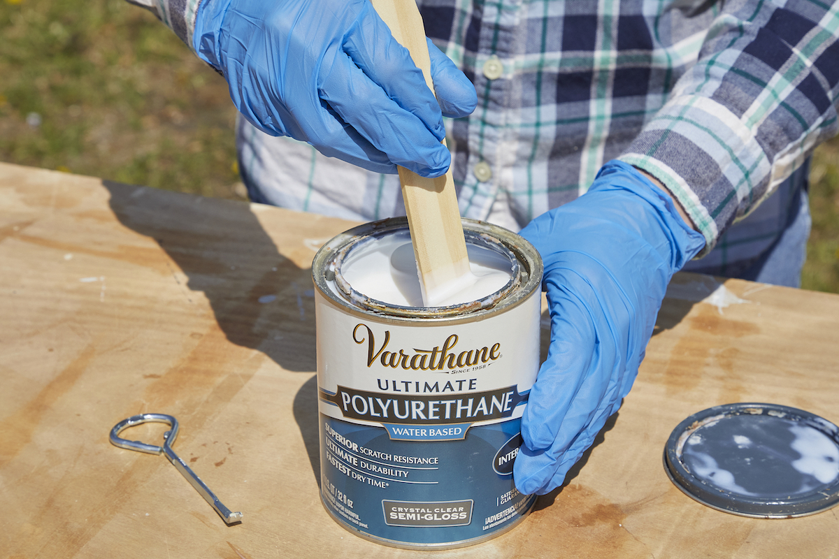 Woman wearing disposable gloves stirs a can of polyurethane with a wooden stir stick.