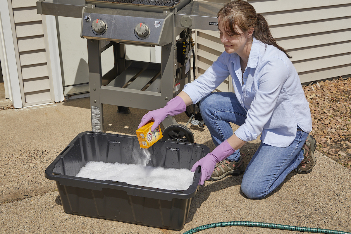 Crouched in front of a gas grill, a woman pours baking soda into a a large plastic basin filled with soapy water.