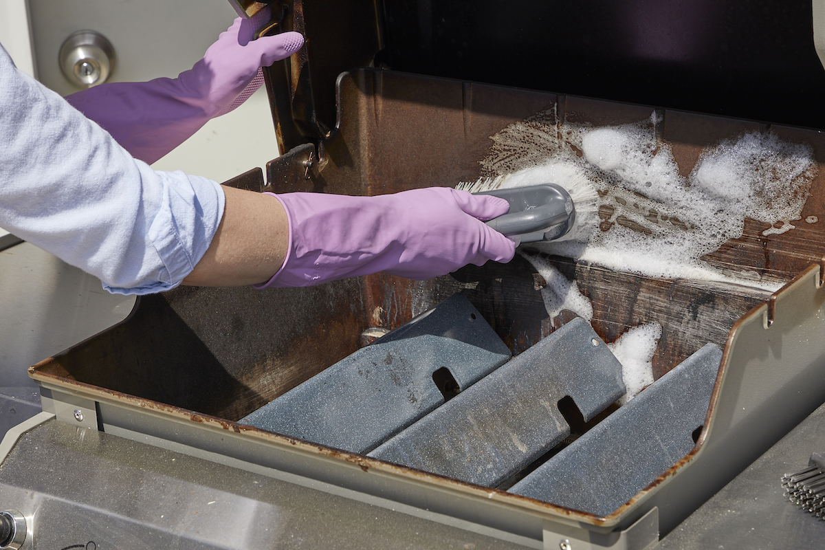 Woman wearing rubber gloves uses a scrub brush to scrub metal interior of gas grill.