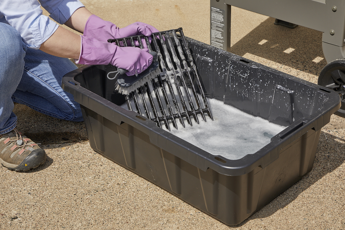 Woman wearing rubber gloves crouches over a plastic bin filled with soapy water, scrubbing grease off a grill grate.
