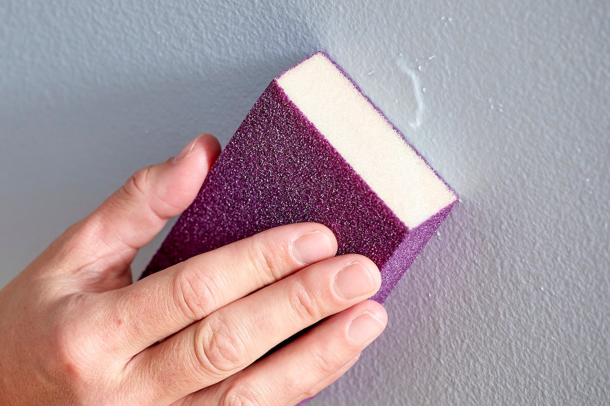 Woman uses a sanding block to sand a patch in drywall.