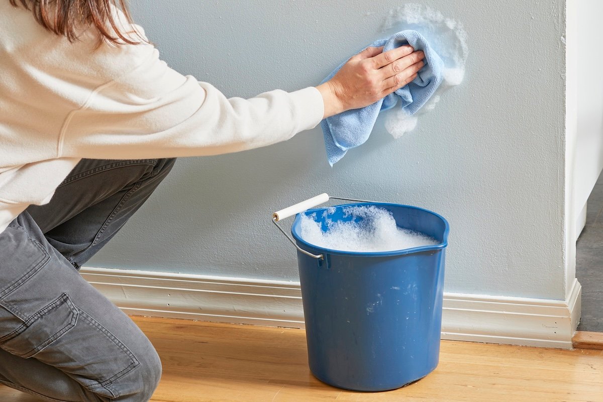 Woman dips rag into a bucket of soapy water and uses rag to wash a wall.