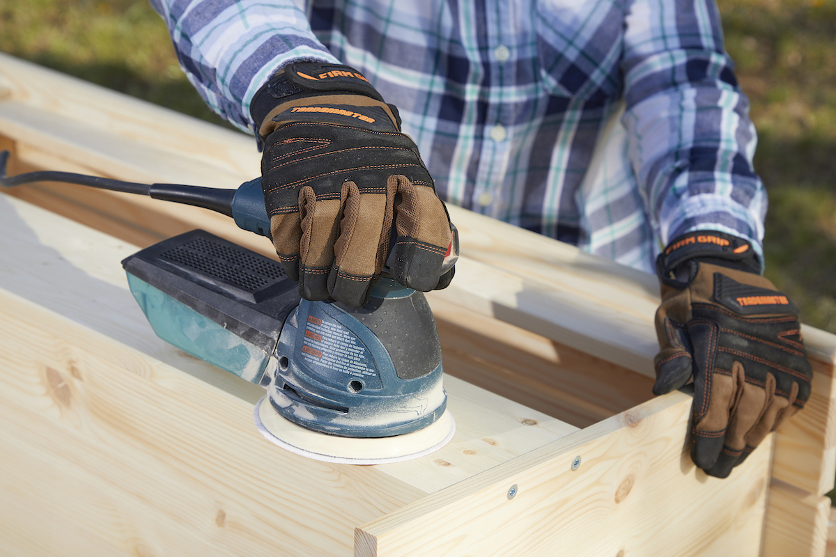 Woman wearing work gloves uses palm sander to sand wood.