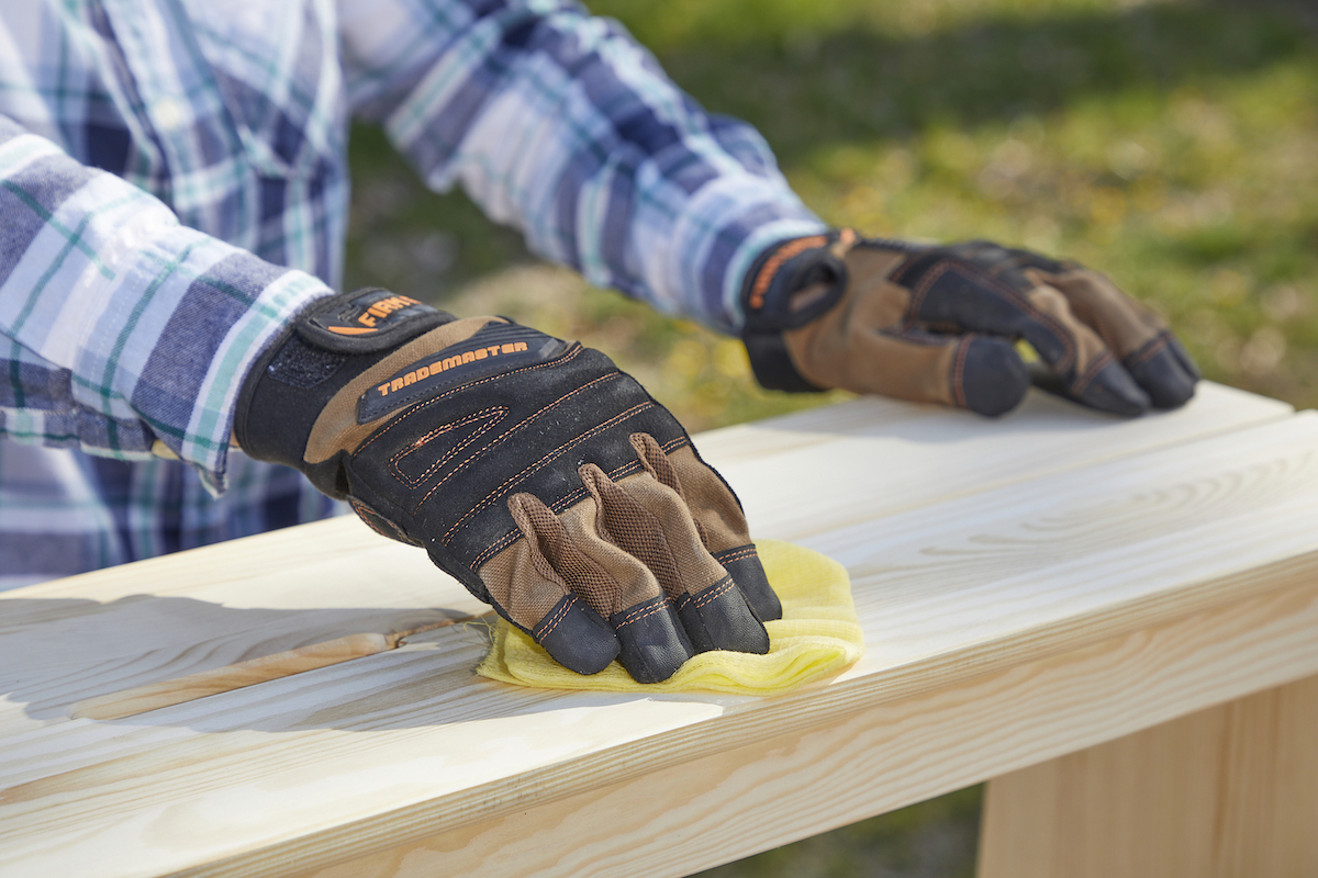 Woman wearing work gloves wipes sanded wood with a yellow cloth.