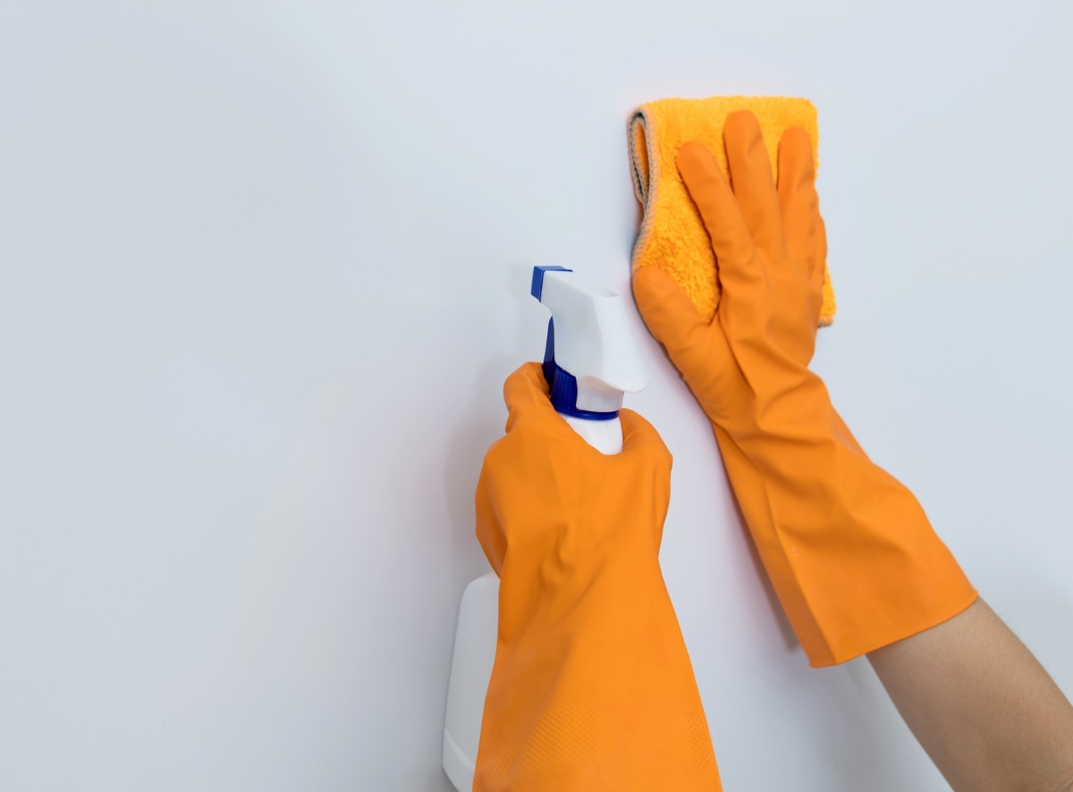Orange gloved hands are cleaning a white wall with a rag and spray cleaner.