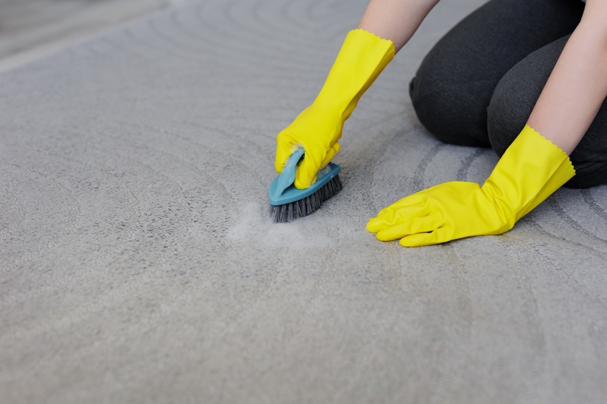 A person wearing yellow rubber gloves scrubs carpet with a soapy solution.