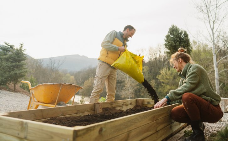 Photo of a couple working on a raised garden bed in their yard by the river. The man pours a bag of soil into the bed, standing next to a wheelbarrow.