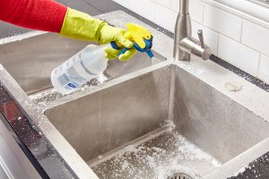 Woman sprays vinegar into a stainless steel sink that has been scrubbed with baking soda.