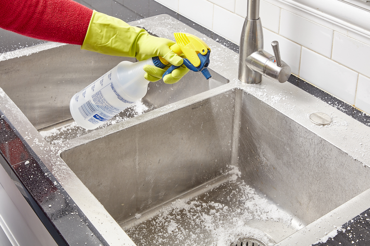 Woman sprays vinegar into a stainless steel sink that has been scrubbed with baking soda.