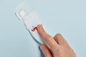 Woman uses a plastic putty knife to scrape excess spackle off a wall.